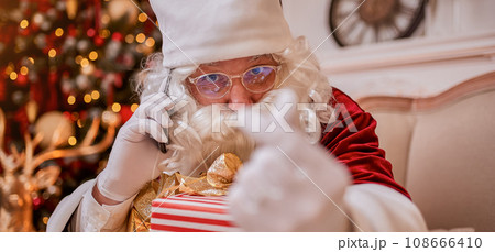 Santa Claus talking on phone sitting neat christmas tree with gifts. New year and Merry Christmas Santa Claus talking on phone sitting neat christmas tree with gifts. New year and Merry Christmas 108666410