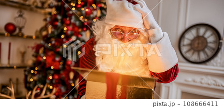 Surprised Santa Claus with glowing gift near christmas tree. New year and Merry Christmas concept Surprised Santa Claus with glowing gift near christmas tree. New year and Merry Christmas concept 108666411