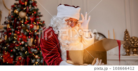 Surprised Santa Claus with glowing gift near christmas tree. New year and Merry Christmas concept Surprised Santa Claus with glowing gift near christmas tree. New year and Merry Christmas concept 108666412