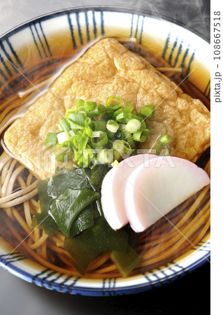 油揚げ、かまぼこ、ワカメ、葱が乗った熱々きつね蕎麦を黒背景で俯瞰撮影 108667518
