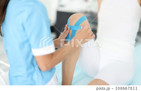 Rehabilitation therapist gluing blue tapes on woman knee closeup Rehabilitation therapist gluing blue tapes on woman knee closeup 108667811