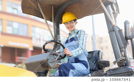 Young woman builder in hard hat sitting behind wheel of asphalt paver Young woman builder in hard hat sitting behind wheel of asphalt paver 108667841