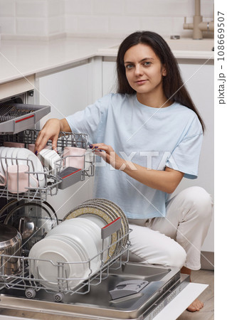 pretty woman unloading from open automatic built-in dishwasher machine with clean utensils inside in modern white kitchen. pretty woman unloading from open automatic built-in dishwasher machine with clean utensils inside in modern white kitchen. 108669507