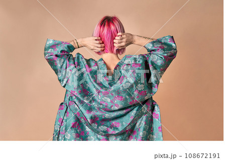 A young girl stands with her back thrown back with her hands behind her head on a light background. A girl with fashionable pink hair, dressed in a blue kimono. Studio shot 108672191