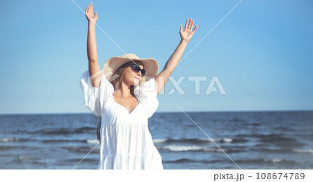 Happy blonde woman is on the ocean beach in a white dress and sunglasses, raising hands Happy blonde woman is on the ocean beach in a white dress and sunglasses, raising hands 108674789