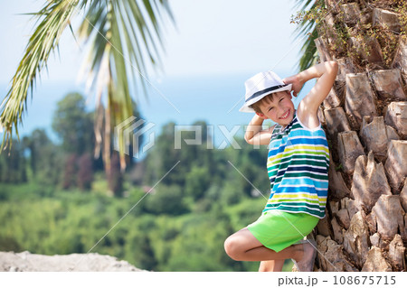 a happy boy in a panama stands near a tropical palm tree, looks at the camera and smiles. A child on vacation in a tropical country. 108675715