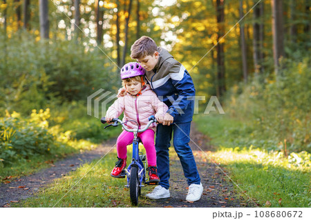 Cute little preschool girl in safety helmet riding bicycle. School kid boy, brother teaching happy healthy sister child cycling and having fun with learning bike. Active siblings family outdoors. Cute little preschool girl in safety helmet riding bicycle. School kid boy, brother teaching happy healthy sister child cycling and having fun with learning bike. Active siblings family outdoors. 108680672
