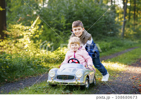 Two happy children playing with big old toy car in autumn forest, outdoors. Kid boy pushing and driving car with little toddler girl, cute sister inside. Laughing and smiling kids. Lovely family 108680673