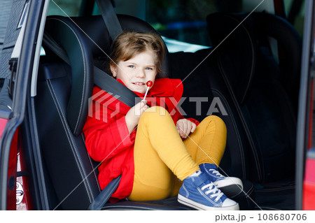 Little toddler girl sitting in car seat, holding sweet lollipop against nausea and sickness and looking out of the window on nature and traffic. Cute kid traveling by car. Child safety on the road. 108680706