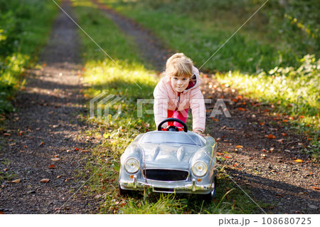 Little preschool girl driving big vintage toy car. Happy child having fun with playing outdoors. Active preschooler child enjoying warm autumn day in forest. Smiling stunning kid playing Little preschool girl driving big vintage toy car. Happy child having fun with playing outdoors. Active preschooler child enjoying warm autumn day in forest. Smiling stunning kid playing 108680725