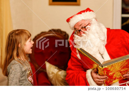 Little toddler girl talking to Santa Claus called Nikolaus or Weihnachtsmann in German. Happy smiling kid waiting for gifts. Santa with golden book. Xmas, childhood, holiday 108680754