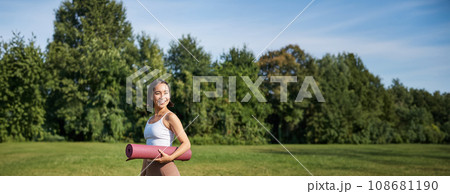 Vertical shot of stylish fitness woman with rubber mat, posing on lawn in park, doing workout on fresh air 108681190