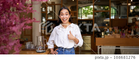 Happy asian woman shows thumbs up, approves, likes smth good, stands near cafe entrance and give her recommendation Happy asian woman shows thumbs up, approves, likes smth good, stands near cafe entrance and give her recommendation 108681276
