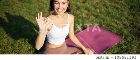 Portrait of asian woman sitting on rubber mat, meditating, showing okay sign and looking at camera, doing yoga Portrait of asian woman sitting on rubber mat, meditating, showing okay sign and looking at camera, doing yoga 108681393