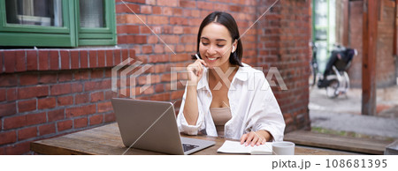 Young smiling asian woman with wireless earphones, sitting in coworking office space with laptop, doing homework, making notes 108681395