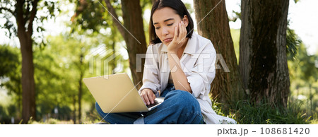 Portrait of asian woman sitting on grass near tree, using laptop, working, doing homework remotely on sunny summer day 108681420