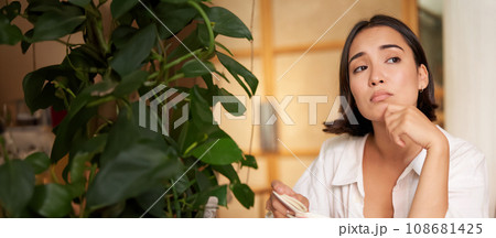 Vertical shot of asian girl sits alone in cafe, reads book and looks upset, drinks coffee with croissant Vertical shot of asian girl sits alone in cafe, reads book and looks upset, drinks coffee with croissant 108681425