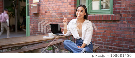 Enthusiastic asian woman, pointing at upper right corner, showing advertisement, news on banner, looking impressed and intrigued by smth 108681456