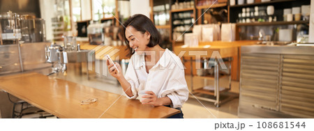 Beautiful asian girl, smiling and reading on smartphone, drinking coffee, sitting in cafe, looking at message on mobile phone Beautiful asian girl, smiling and reading on smartphone, drinking coffee, sitting in cafe, looking at message on mobile phone 108681544