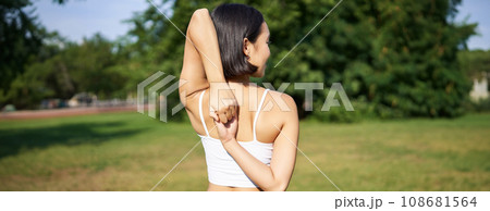 Rear view of young sporty woman stretching her arms behind back, warm-up, prepare for workout jogging, sport event in park Rear view of young sporty woman stretching her arms behind back, warm-up, prepare for workout jogging, sport event in park 108681564
