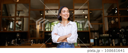 Portrait of confident young asian woman, business owner, cross arms on chest, looking pleased, standing in front of cafe 108681641