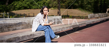 Depressed asian girl sits on bench in park with smartphone, feeling uneasy and stressed, frowning and sighing 108681821