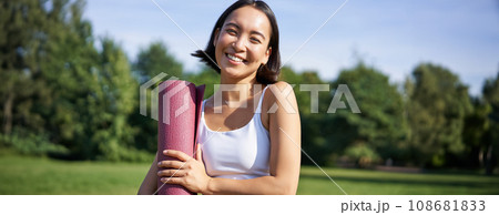 Portrait of asian girl smiles and laughs, stands with sports equipment, rubber mat, wears uniform for workout and exercises in park Portrait of asian girl smiles and laughs, stands with sports equipment, rubber mat, wears uniform for workout and exercises in park 108681833