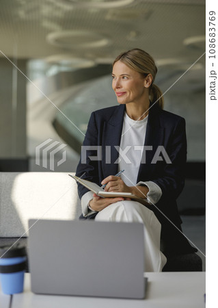 Confident female manager making notes in note pad sitting on modern office background Confident female manager making notes in note pad sitting on modern office background 108683769