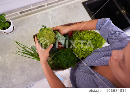 View from above of woman unpacking vegetables, standing by kitchen counter. Healthy eating. Dieting and slimming concept View from above of woman unpacking vegetables, standing by kitchen counter. Healthy eating. Dieting and slimming concept 108684852