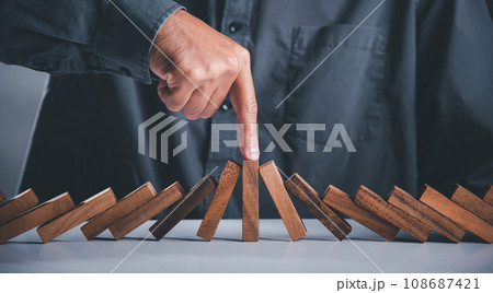 Close up hand stopping or preventing falling block in line with risk project control, Finger of businessman pointing one wooden to stopping domino effect stopped, Risk protection concept 108687421