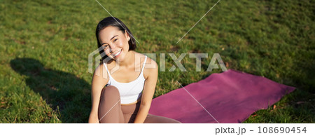 Vertical shot of smiling asian girl meditating, sitting on yoga mat with smartphone, doing yoga on fresh air in park, watching online fitness instructor Vertical shot of smiling asian girl meditating, sitting on yoga mat with smartphone, doing yoga on fresh air in park, watching online fitness instructor 108690454