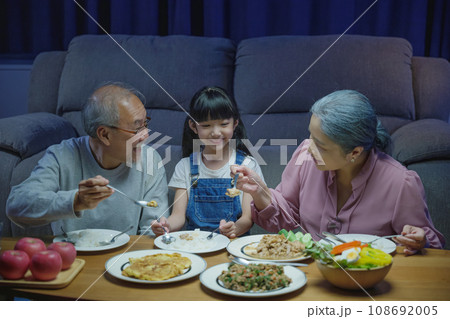 Happy Asian senior parent and child eating dinner food together in living room indoors, family grandmother grandfather and granddaughter dining on table and having fun during at home night time 108692005
