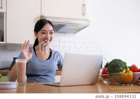 Happy woman asian in stylish tracksuit fresh smoothie shooting video with modern laptop in kitchen Happy woman asian in stylish tracksuit fresh smoothie shooting video with modern laptop in kitchen 108694132