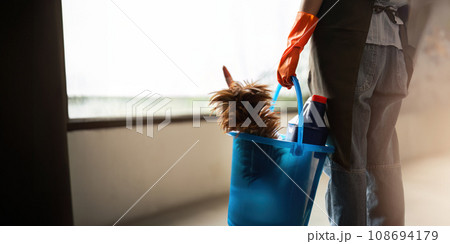 A cleaning woman is standing inside a building holding a blue bucket fulfilled with chemicals and facilities for tidying up in her hand A cleaning woman is standing inside a building holding a blue bucket fulfilled with chemicals and facilities for tidying up in her hand 108694179