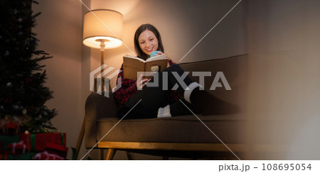 Smiling woman with a cup of tea and a book relaxing in side of a decorated Christmas tree in living room. concept holidays Christmas festive celebrate and tree Xmas seasonal 108695054
