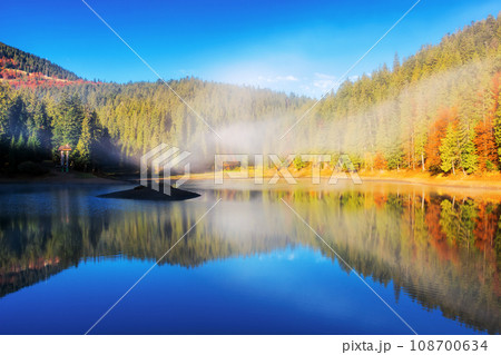 sunny landscape with lake in autumn. fog above the water surface. colorful scenery with sky and trees reflection 108700634