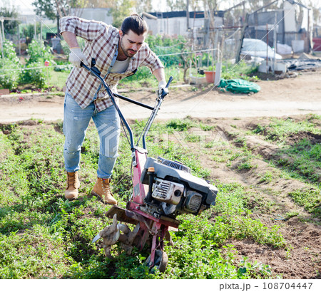 Man professional gardener using plow at land with green grass in garden Man professional gardener using plow at land with green grass in garden 108704447