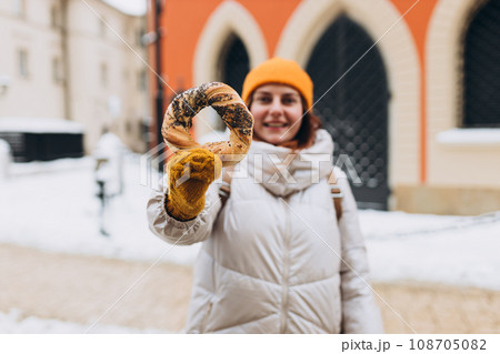 Attractive young female tourist in yellow hat is holding pretzel, traditional polish bagel on Market Square in Krakow. Traveling Europe in winter time. High quality photo 108705082