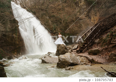 Happy woman in a white dress stands on a stone with a waterfall behind 108705783