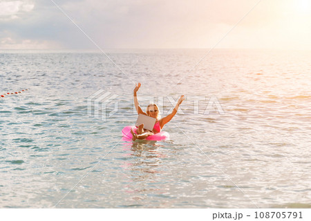 Woman works on laptop in sea. Freelancer, blond woman in sunglases floating on an inflatable big pink donut with a laptop in the sea at sunset. People summer vacation rest lifestyle concept. 108705791