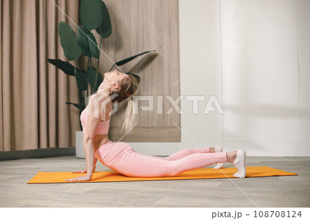 Sporty young woman training at home. Woman wearing rose top and leggins. Woman doing yoga on an orange mat. Sporty young woman training at home. Woman wearing rose top and leggins. Woman doing yoga on an orange mat. 108708124