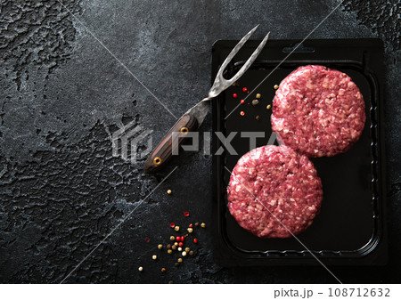 Raw minced beef burgers in plastic vacuum tray on black stone background with fork. 108712632