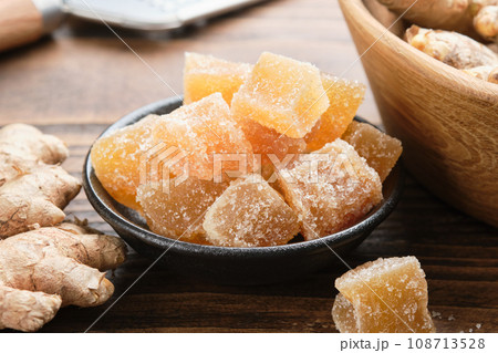 Bowl of candied ginger pieces closeup and ginger roots on table. Healthy sweets. Bowl of candied ginger pieces closeup and ginger roots on table. Healthy sweets. 108713528