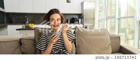 Portrait of cute young woman eating breakfast, having cereals with milk on sofa in living room, smiling and looking satisfied 108713741