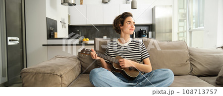 Portrait of girl resting in living room, playing ukulele, learns how to play new musical instrument, spending time relaxing at home 108713757