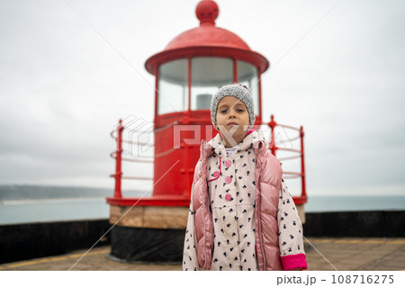 Little girl stands near red lighthouse on rocky seashore 108716275