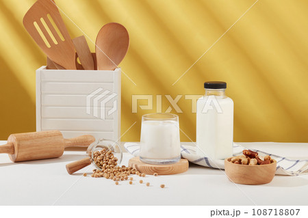 Front view of a milk cup placed on wooden podium, displayed with a bowl of nuts, soybeans, kitchen utensil and a milk bottle. Empty label bottle for product promotion 108718807