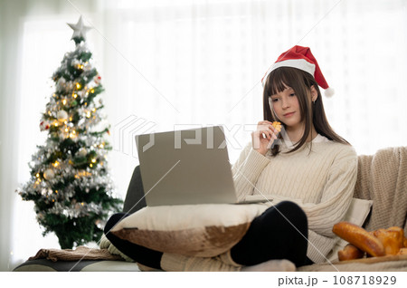 A pretty woman in a Santa hat is snacking and watching online videos on her laptop on a couch. 108718929