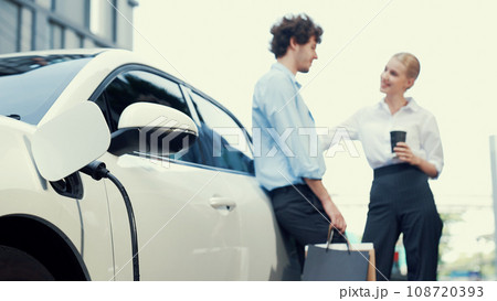 Progressive businessman and businesswoman leaning on EV car and charging point. Progressive businessman and businesswoman leaning on EV car and charging point. 108720393