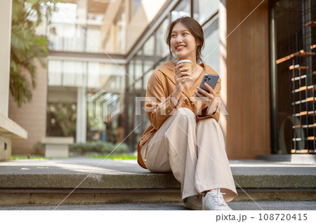 Beautiful Asian woman is sitting on the stairs with her phone and a takeaway coffee cup in her hands Beautiful Asian woman is sitting on the stairs with her phone and a takeaway coffee cup in her hands 108720415